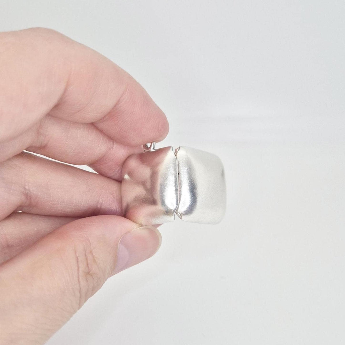A hand holds the sterling silver square locket pendant in a closed position, viewed from the top, emphasizing its thickness and the seam where it closes.