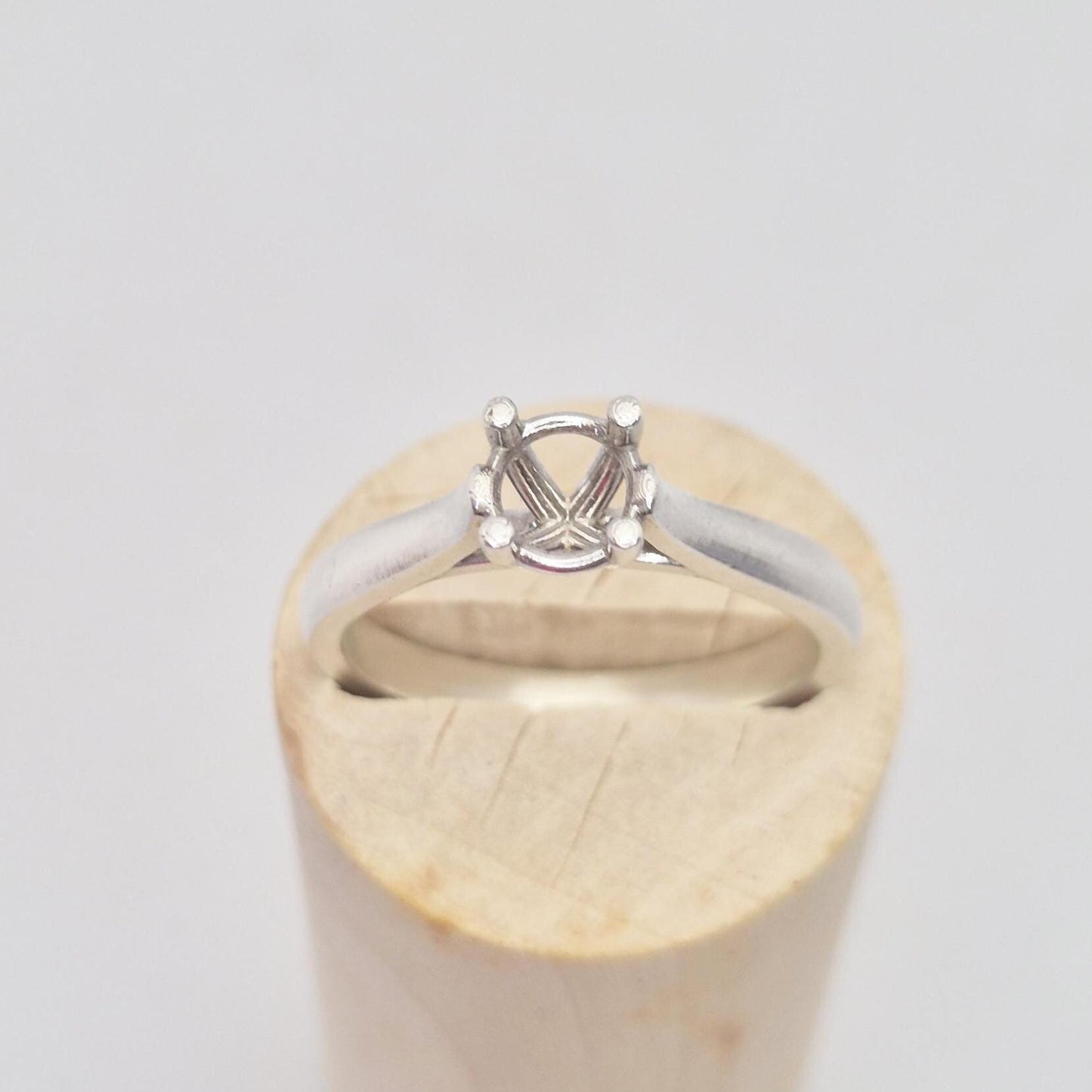 Frontal close-up of a sterling silver ring setting with a four-prong round head, presented on a cylindrical wooden stand, focusing on the details of the stone mounting.