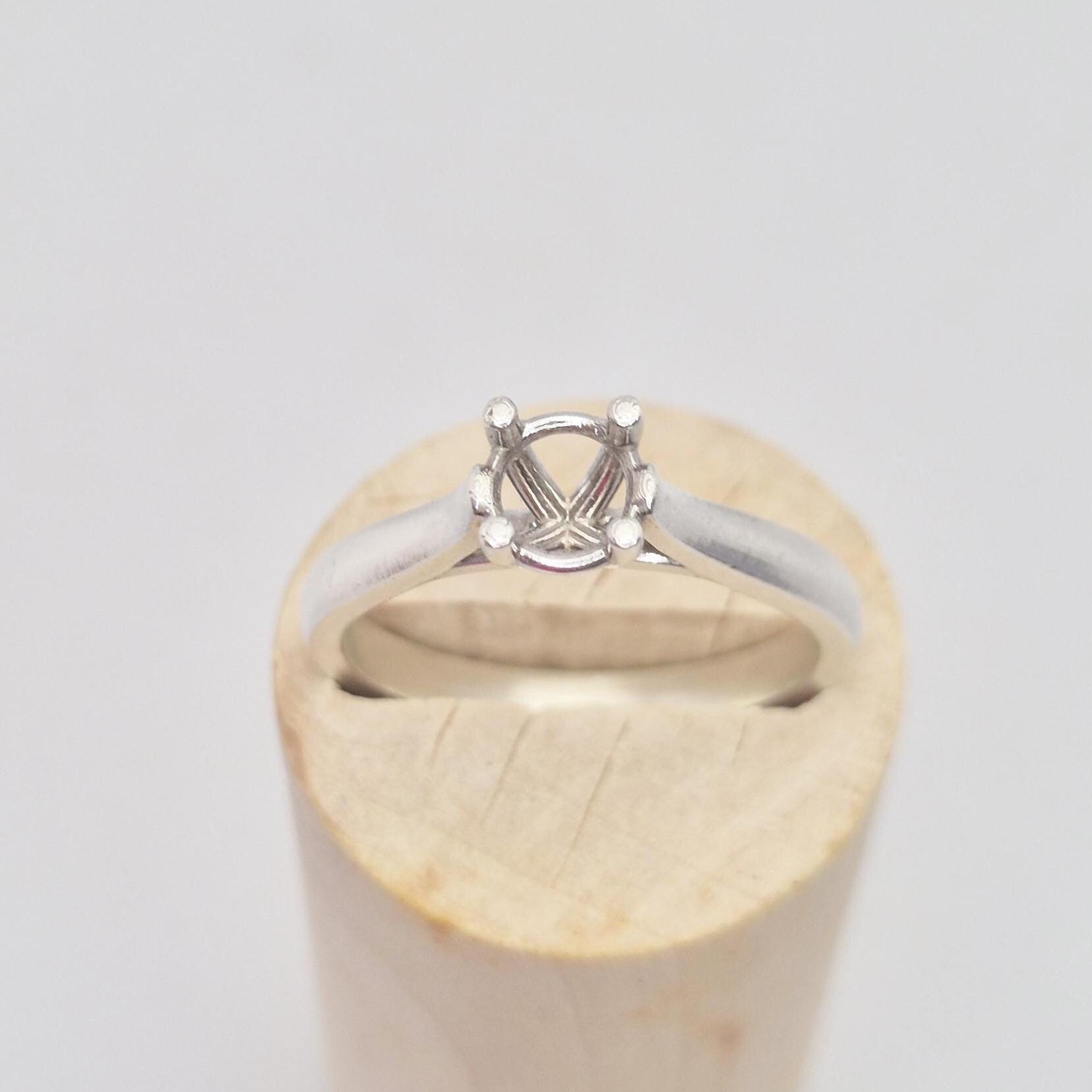 Frontal close-up of a sterling silver ring setting with a four-prong round head, presented on a cylindrical wooden stand, focusing on the details of the stone mounting.