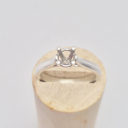 Frontal close-up of a sterling silver ring setting with a four-prong round head, presented on a cylindrical wooden stand, focusing on the details of the stone mounting.