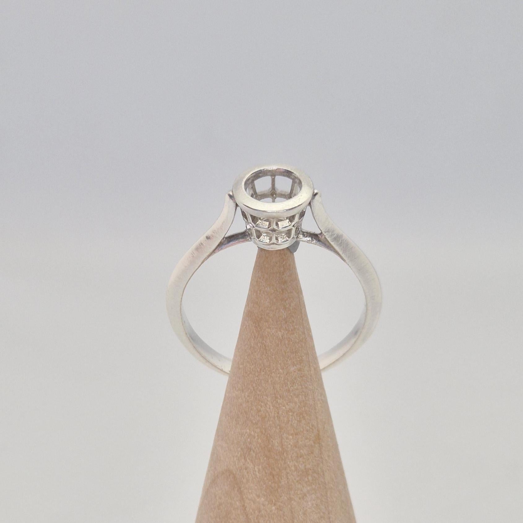 Front view of a silver ring setting with an empty round bezel, displayed upright on a light wooden cone. The setting has an open basket design beneath the bezel.