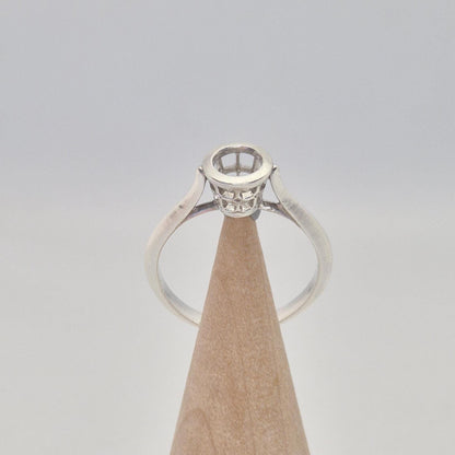 Front view of a silver ring setting with an empty round bezel, displayed upright on a light wooden cone. The setting has an open basket design beneath the bezel.
