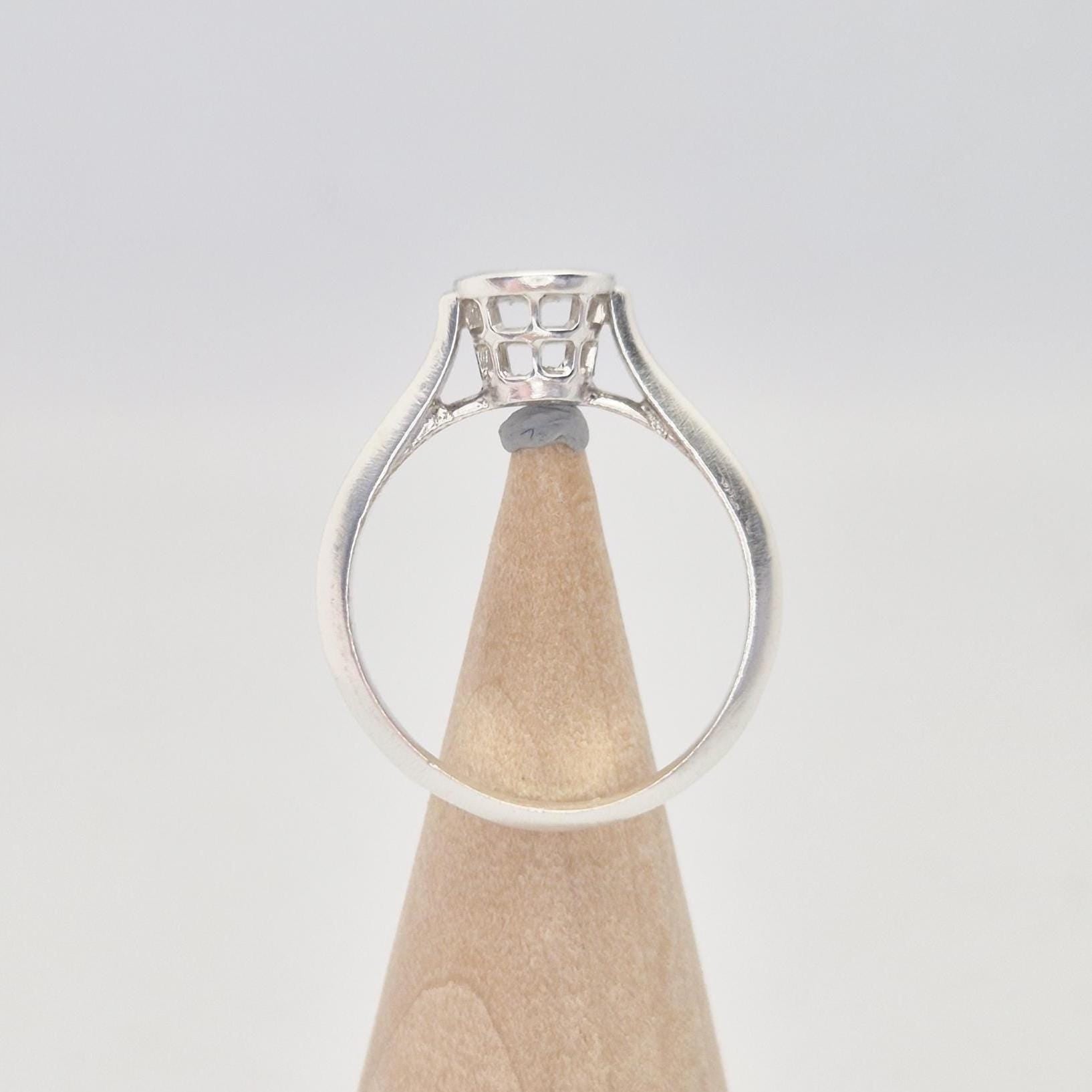 Side view of a silver ring setting with an empty round bezel, displayed upright on a light wooden cone. The ring band is smooth and tapers slightly towards the setting.