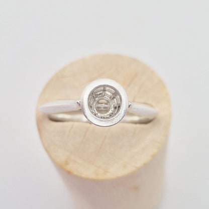 Front-side view of a silver ring setting with an empty round bezel, displayed upright on a light wooden cylinder. The bezel setting is clearly visible.