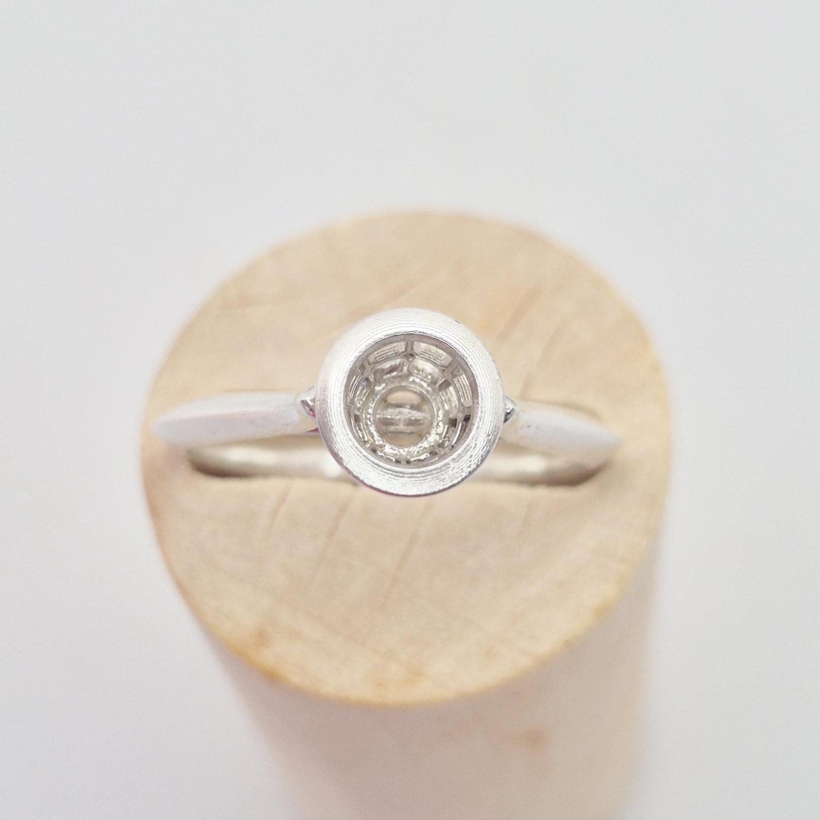 Front-side view of a silver ring setting with an empty round bezel, displayed upright on a light wooden cylinder. The bezel setting is clearly visible.