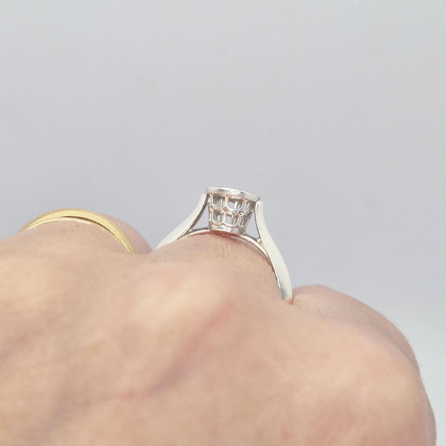 Close-up of a hand wearing a silver ring setting with an empty round bezel, viewed from the side. The ring is positioned on the finger, highlighting its profile and basket design.