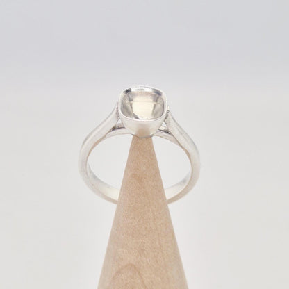 Front view of a sterling silver ring setting with a cushion cut bezel, standing on a cylindrical wooden stand, showing the graceful curve of the band leading to the setting.