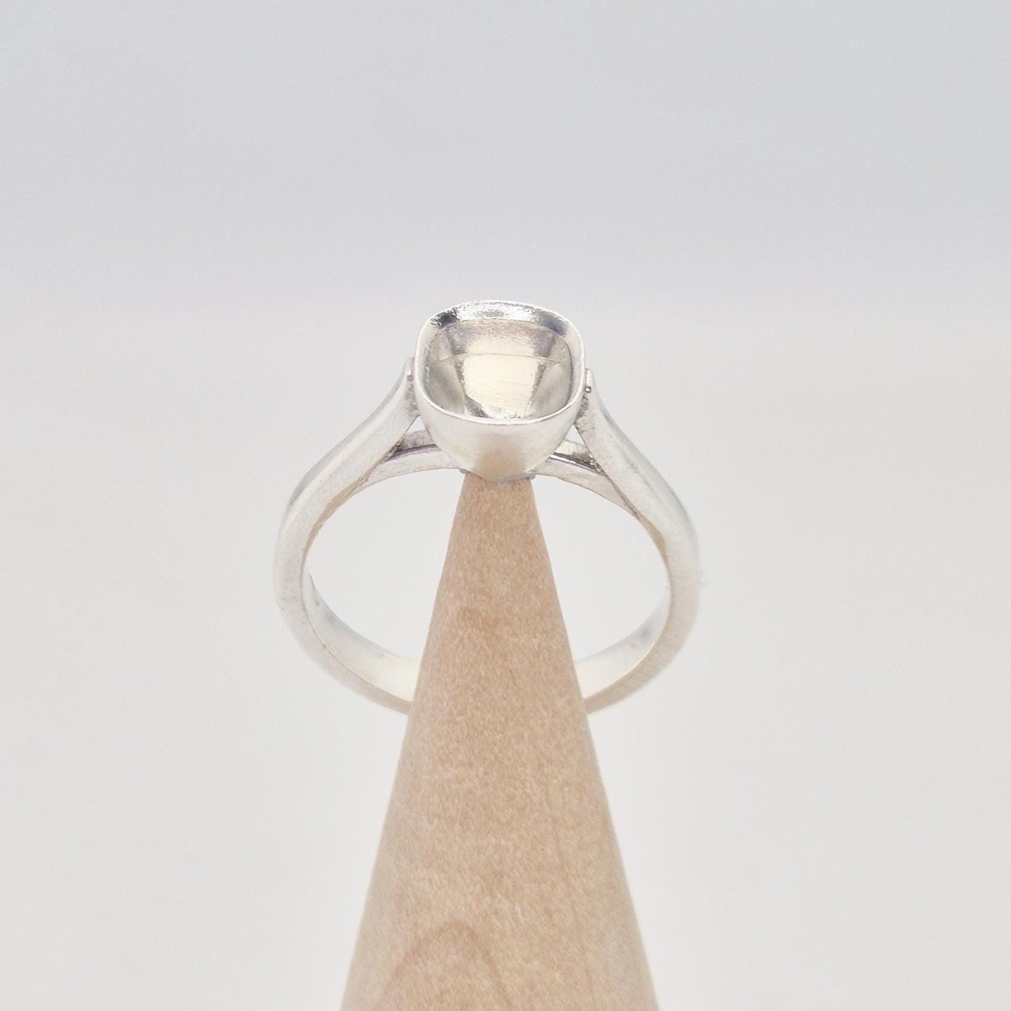 Front view of a sterling silver ring setting with a cushion cut bezel, standing on a cylindrical wooden stand, showing the graceful curve of the band leading to the setting.