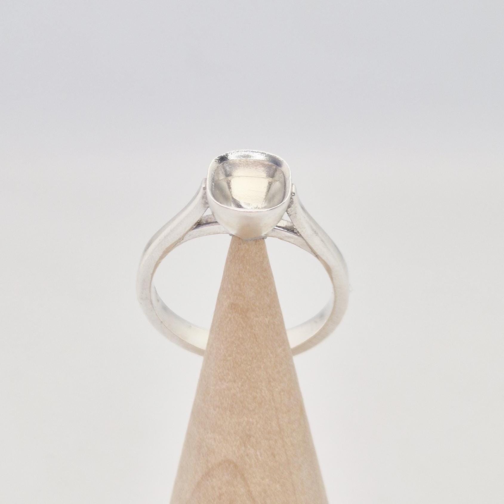 Front view of a sterling silver ring setting with a cushion cut bezel, standing on a cylindrical wooden stand, showing the graceful curve of the band leading to the setting.