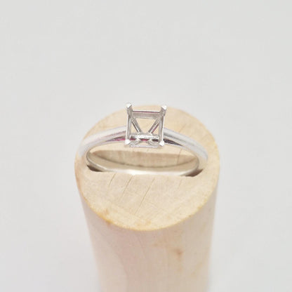 Silver cathedral setting princess cut ring blank resting on a cylindrical wooden ring holder, viewed slightly from the side, showing the empty four-prong setting and the curvature of the band.