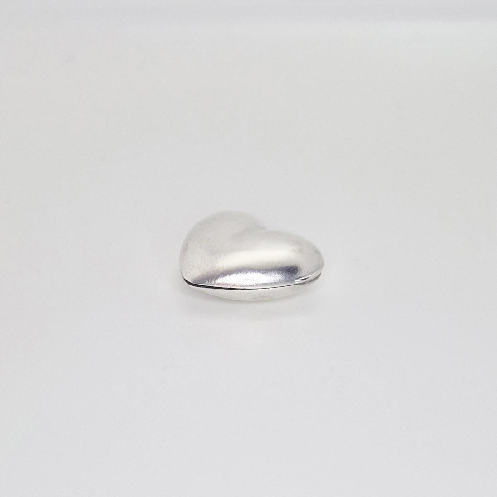 A closed, puffed silver heart locket seen from the back, showing the seam where the two halves join and its rounded contours on a plain white background.
