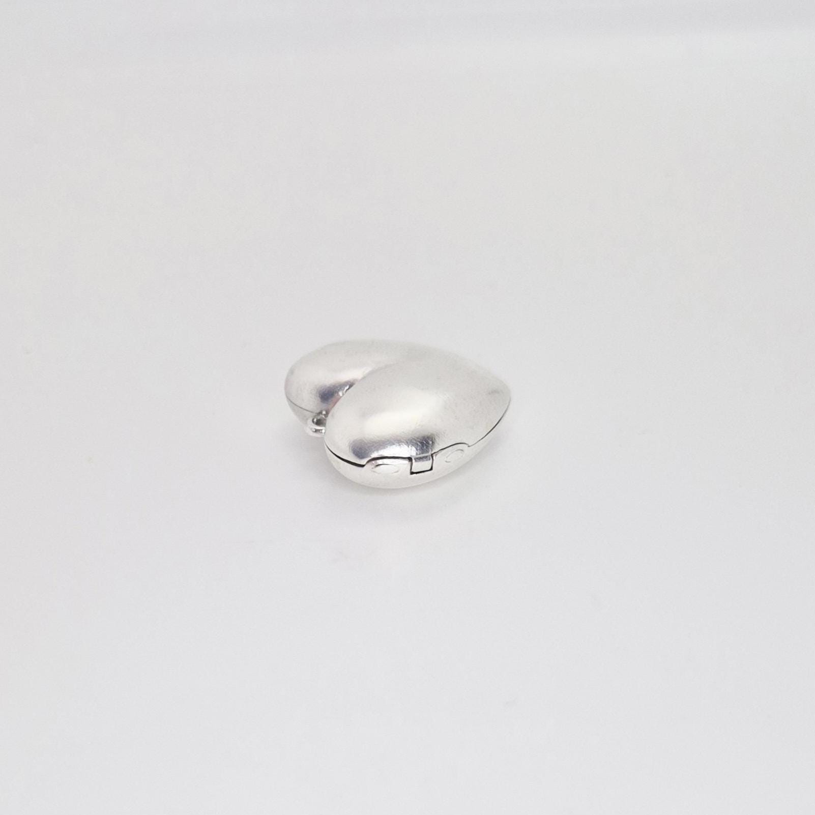 A closed, puffed silver heart locket viewed from the side on a white background, highlighting its thick, rounded profile and the hinge mechanism.