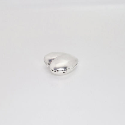A closed, puffed silver heart locket viewed from the side on a white background, highlighting its thick, rounded profile and the hinge mechanism.