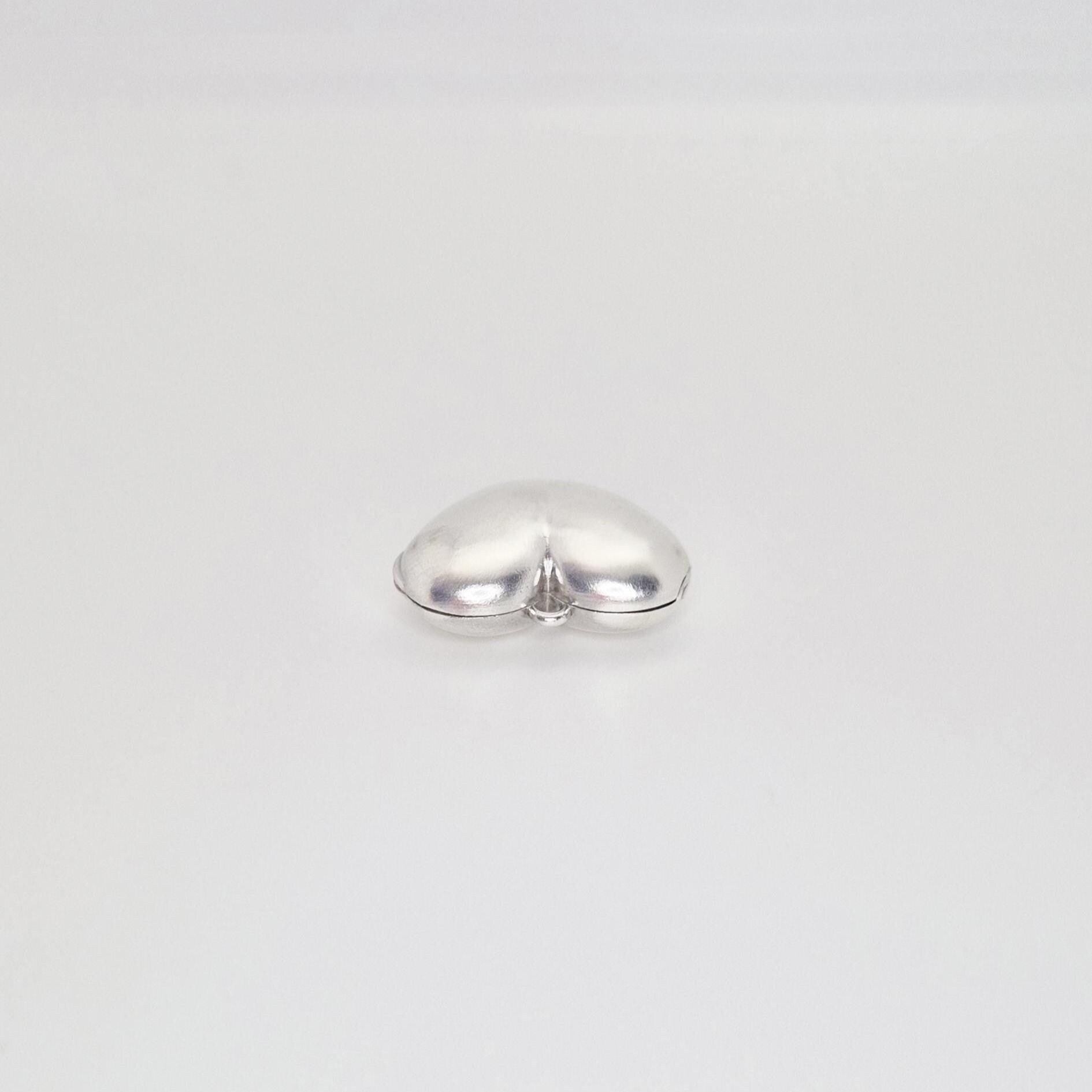 A shiny, puffed silver heart locket, closed and lying on a plain white background, shown from a slightly elevated angle.