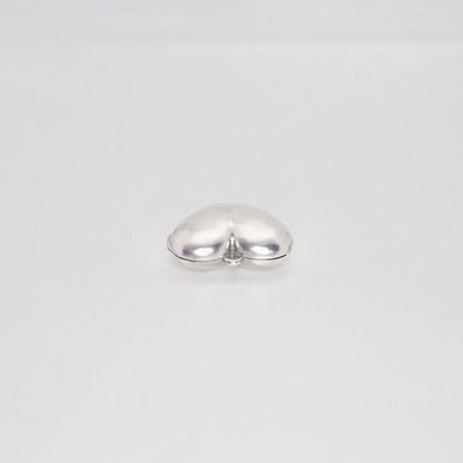 A shiny, puffed silver heart locket, closed and lying on a plain white background, shown from a slightly elevated angle.