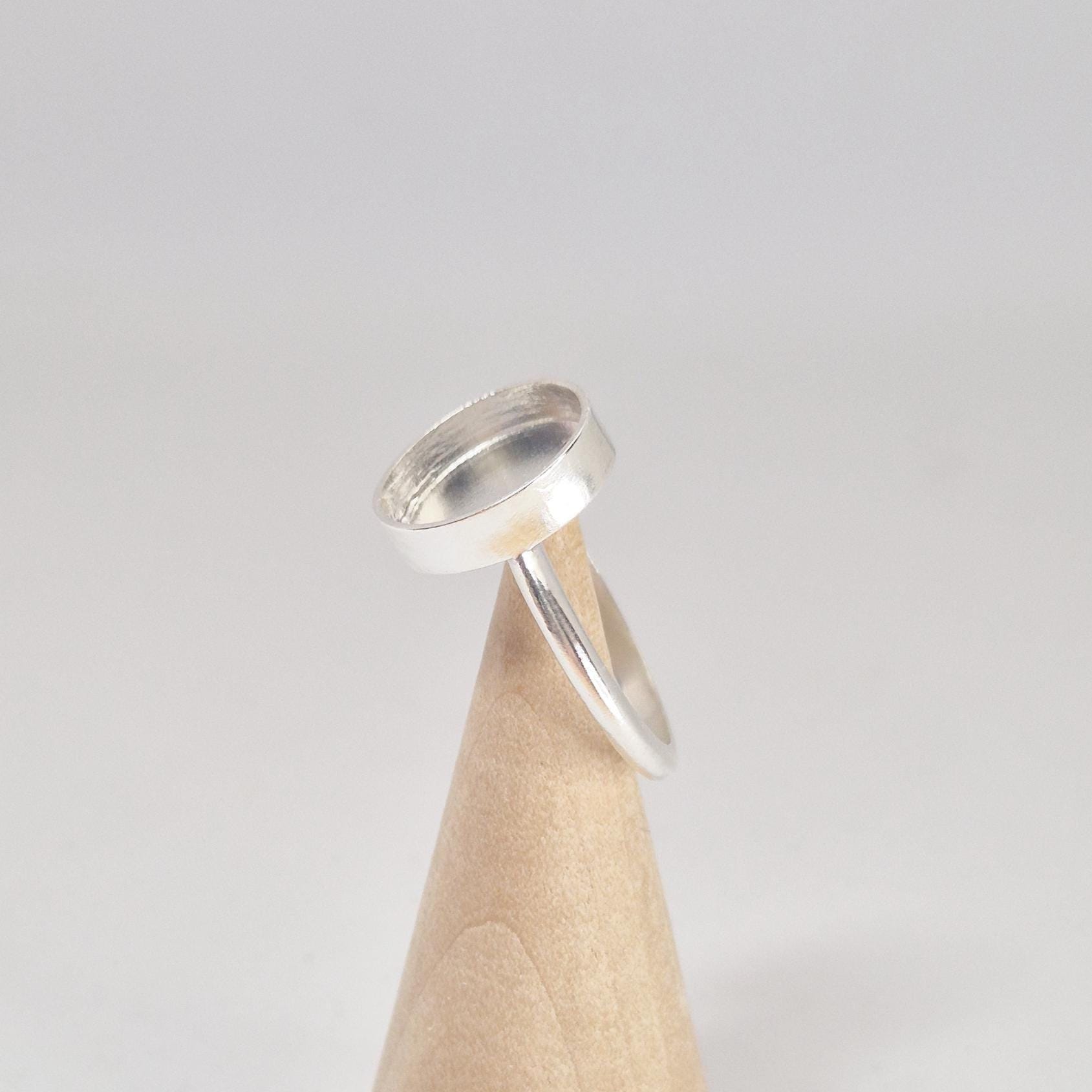 Angled side view of an empty sterling silver oval cabochon ring setting, displayed on a light wood conical stand.