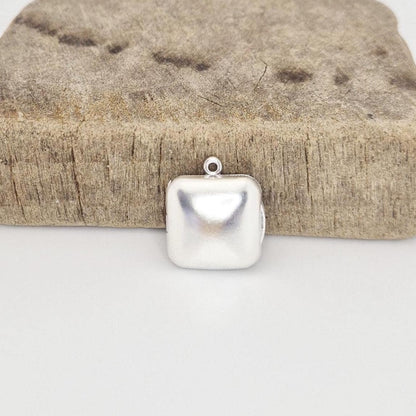 A closed sterling silver square locket pendant with a round bail, positioned in front of a rustic, textured wood block