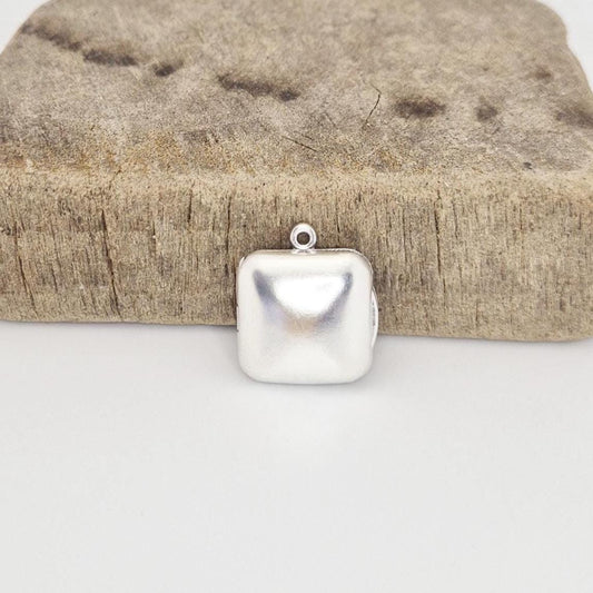 A closed sterling silver square locket pendant with a round bail, positioned in front of a rustic, textured wood block