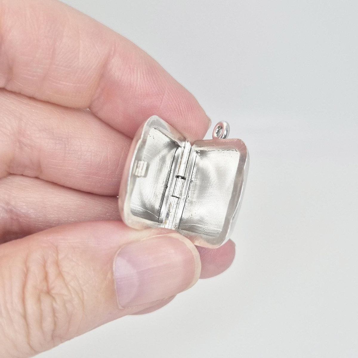 A hand holds an open sterling silver square locket pendant, allowing a clear view into its two internal compartments and the central hinge mechanism.
