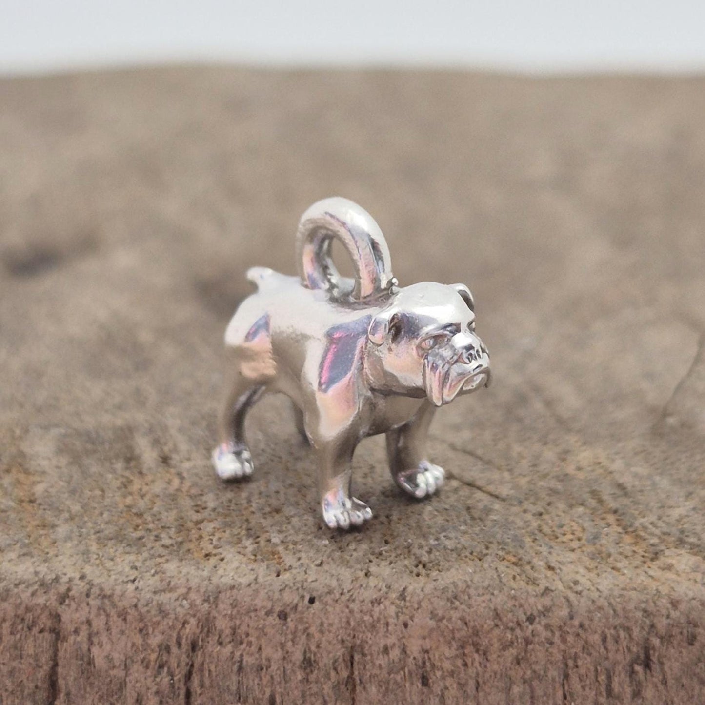 Angled side view of a silver bulldog charm on wood, showing the body shape and rounded top loop for attaching to chains or bracelets.