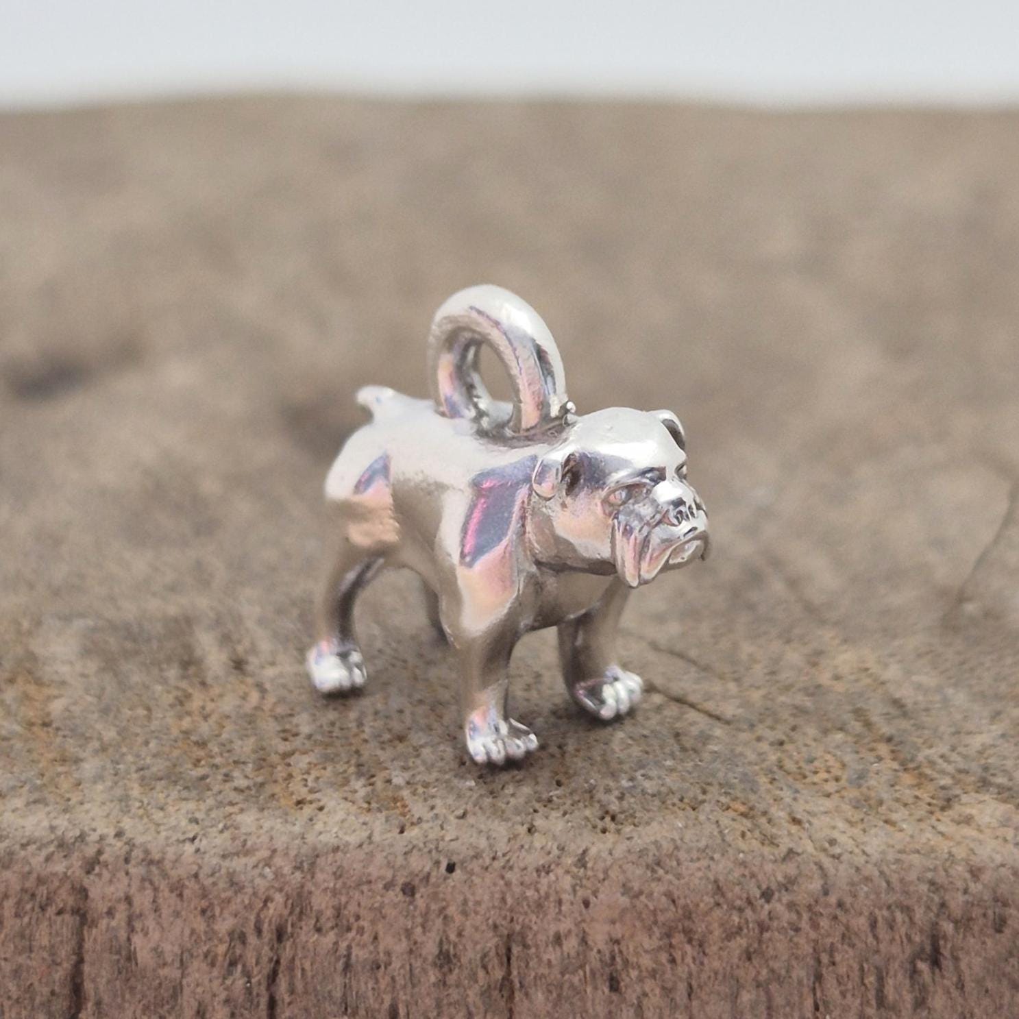 Angled side view of a silver bulldog charm on wood, showing the body shape and rounded top loop for attaching to chains or bracelets.