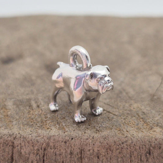Angled side view of a silver bulldog charm on wood, showing the body shape and rounded top loop for attaching to chains or bracelets.