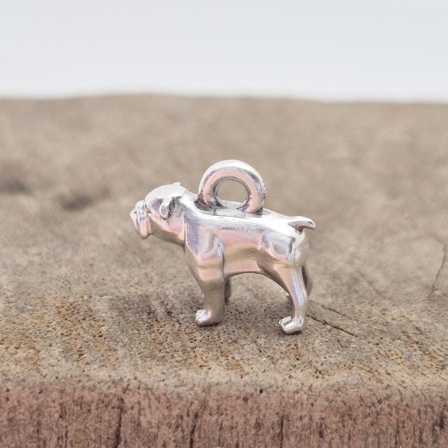 Profile view of a silver bulldog charm standing on a wooden surface, showing leg stance and solid cast form.