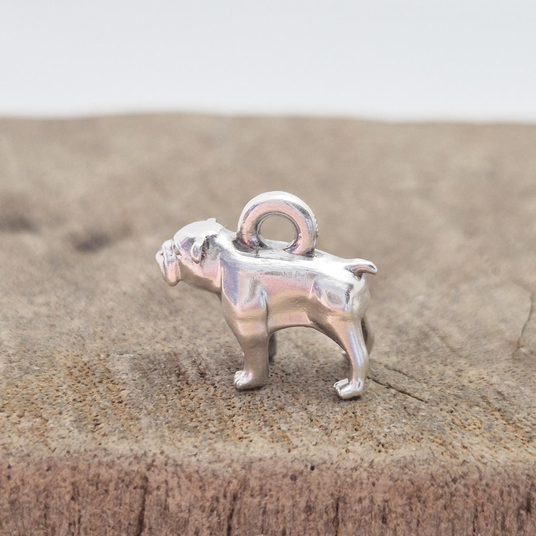 Profile view of a silver bulldog charm standing on a wooden surface, showing leg stance and solid cast form.