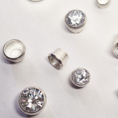 Assorted sterling silver round bezel settings with clear round gemstones on white background, showing unfinished bezel cups for jewellery making.