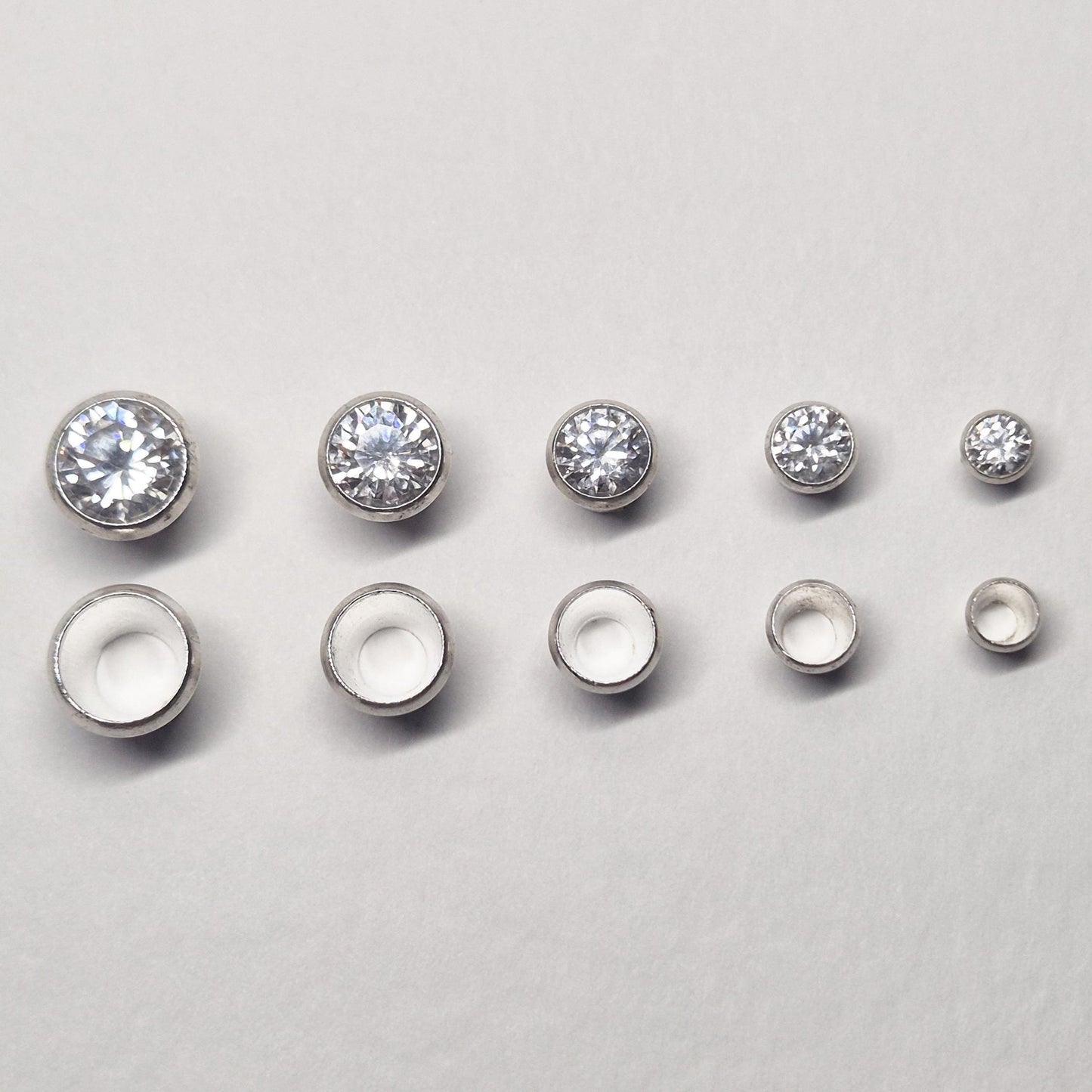 Row of sterling silver bezel cups and matching round stones in multiple sizes, showing graduated bezel settings for jewellery making.