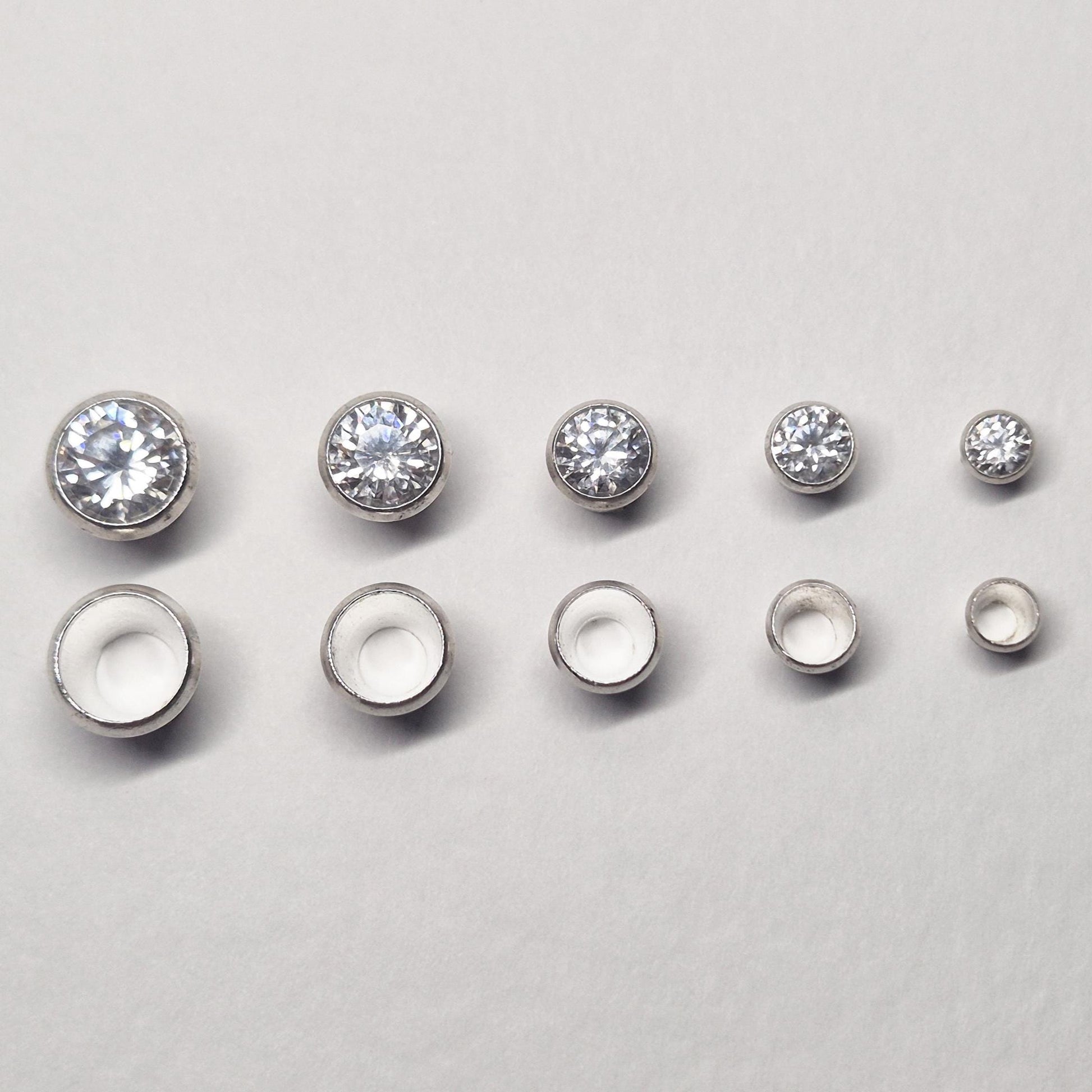 Row of sterling silver bezel cups and matching round stones in multiple sizes, showing graduated bezel settings for jewellery making.