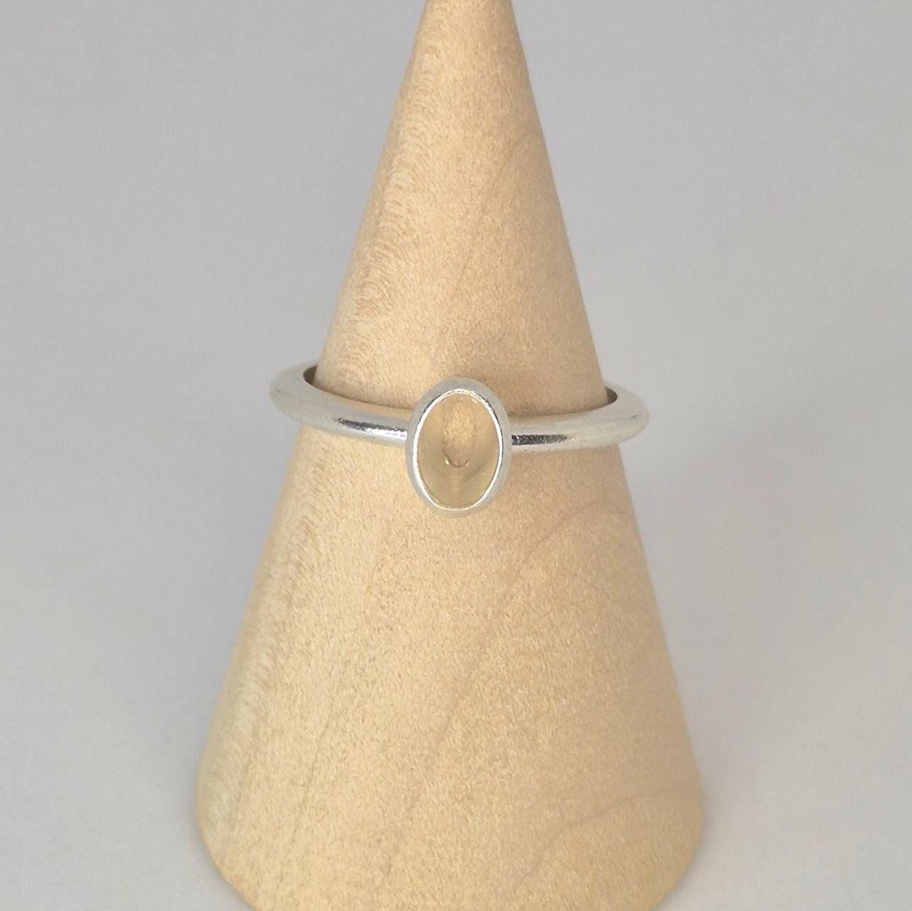 Front view of a sterling or argentium silver 6x4mm Oval Bezel Ring on the middle of a wooden display cone against a white background.