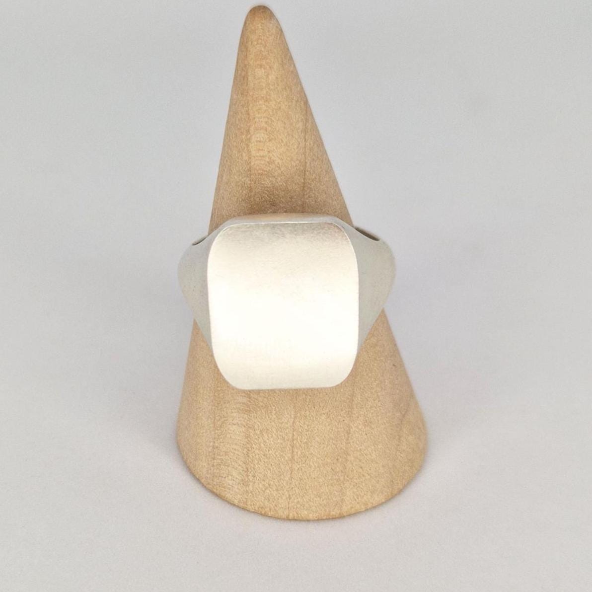 Cushion signet ring displayed on a wooden cone ring stand, featuring a flat 18x16mm rectangular face and matte brushed finish.
