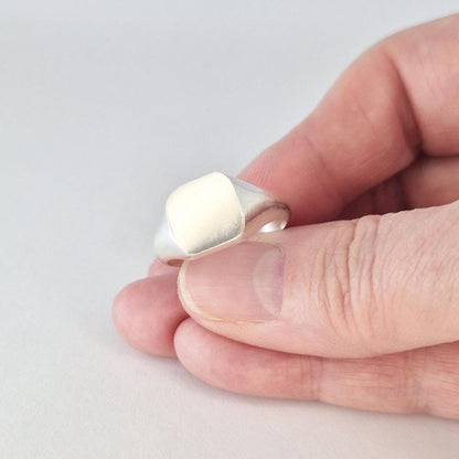 Hand holding small silver cushion signet ring showing scale and polished finish