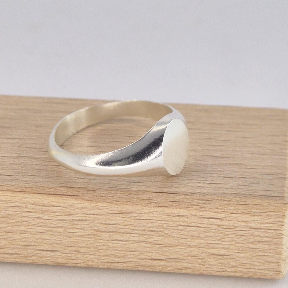 Oval signet ring in polished silver displayed on a wooden block, side profile showing tapered band and smooth oval face