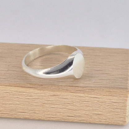 Oval signet ring in polished silver displayed on a wooden block, side profile showing tapered band and smooth oval face