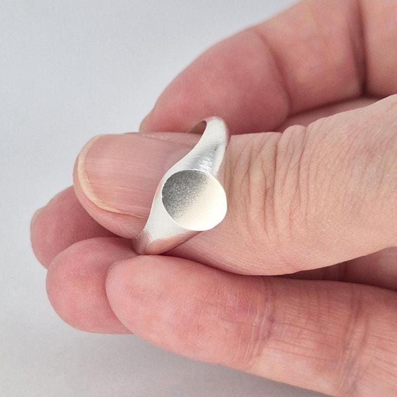 Close-up of oval signet ring held between fingers, showing satin-finish face and polished shoulders