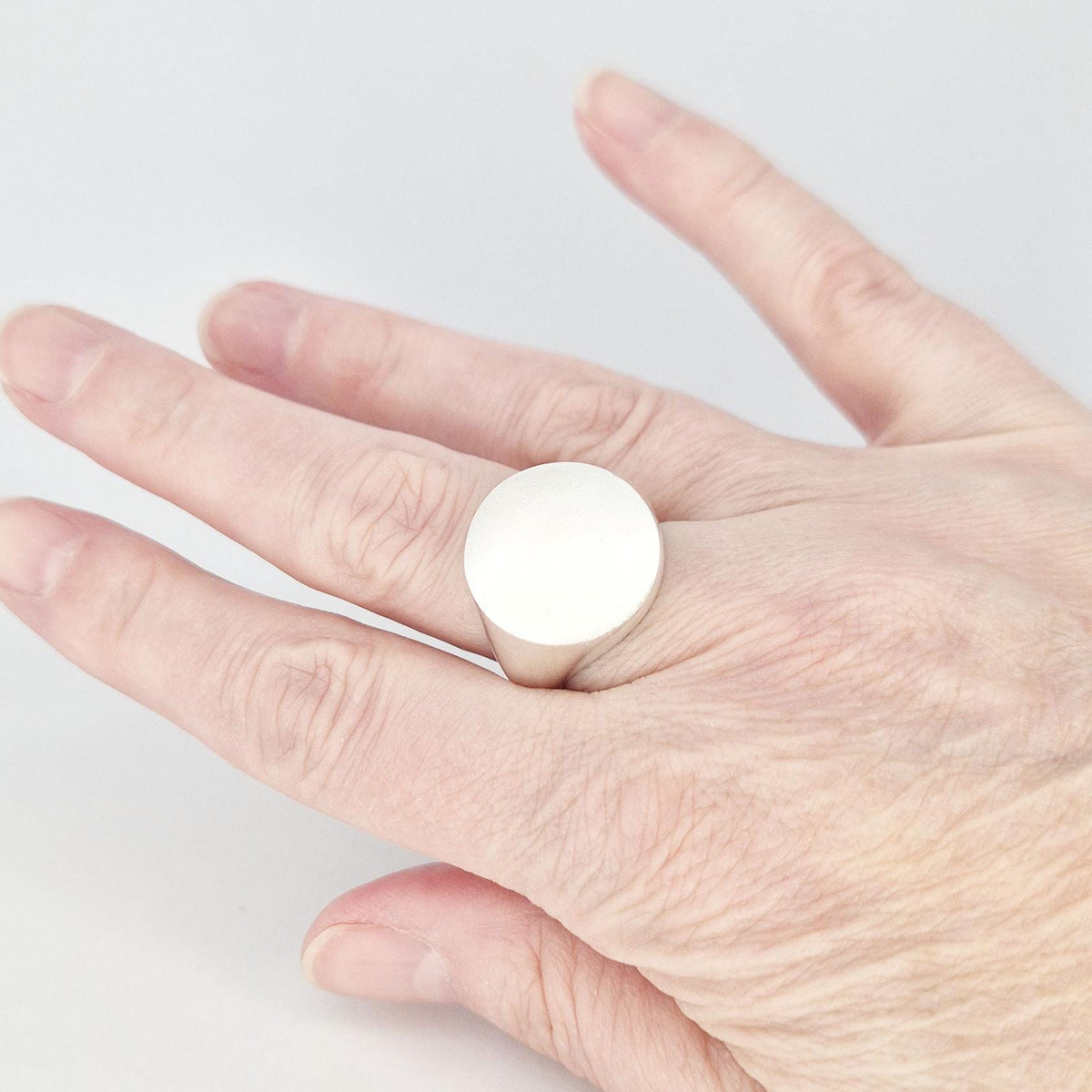 Minimalist round signet ring worn on finger, featuring a large flat circular face in matte silver finish.