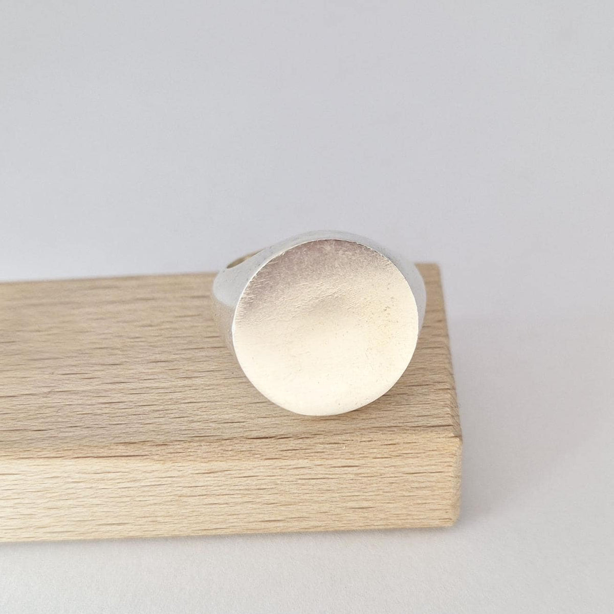 Heavy round signet ring resting on wooden block, showcasing large flat disc face and soft matte silver finish.