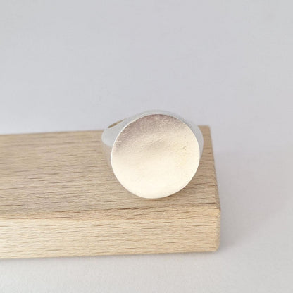 Heavy round signet ring resting on wooden block, showcasing large flat disc face and soft matte silver finish.