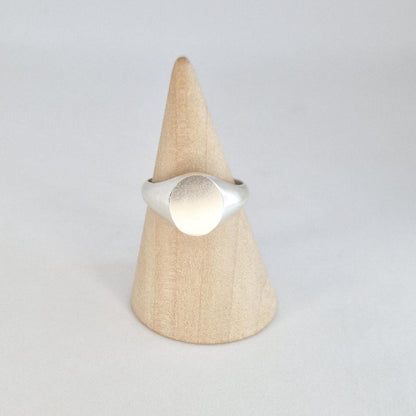 Front-facing oval signet ring on a wooden cone stand with brushed silver face and sleek modern profile.