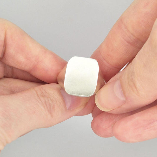 Matte finish cushion signet ring held between fingers, showing the smooth rectangular face with softly rounded corners in sterling or Argentium silver.