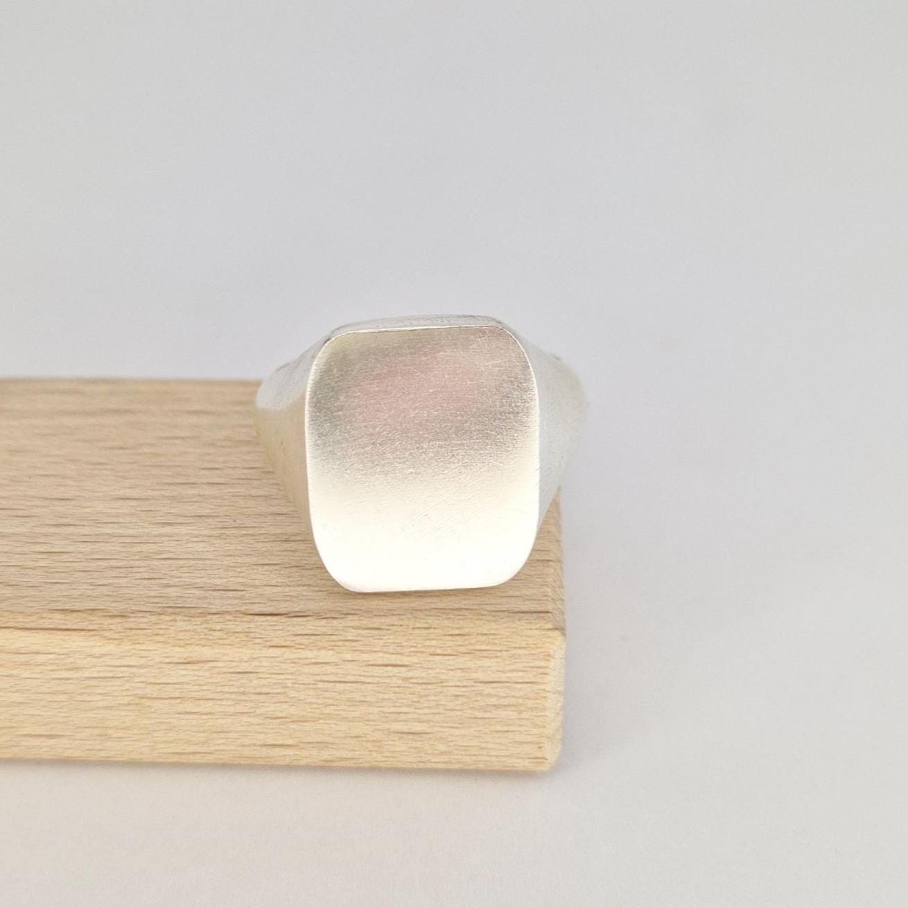 Front view of a blank cushion signet ring resting on a wooden jewellery display stand with brushed silver finish.