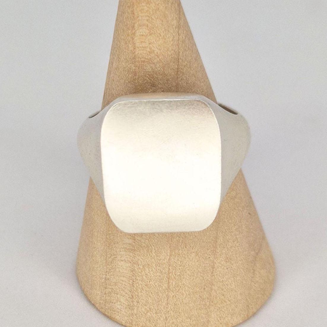 Close-up product photo of a handmade cushion signet ring on wooden cone display, featuring a brushed rectangular face and smooth silver shoulders.